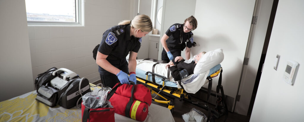 Paramedic students practice emergency response with a patient on a stretcher in a simulation lab.