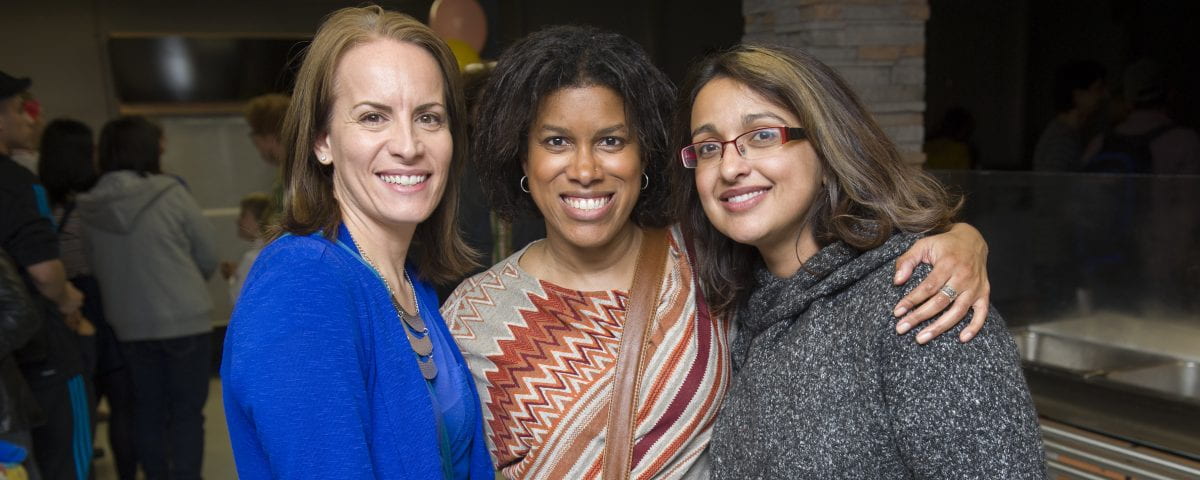 Three women are standing shoulder-to-shoulder while smiling