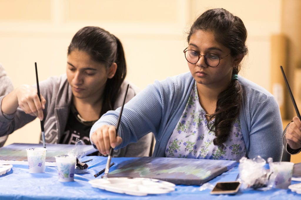 Two students each looking down at a painted canvas while holding a paint brush. The students look relaxed.