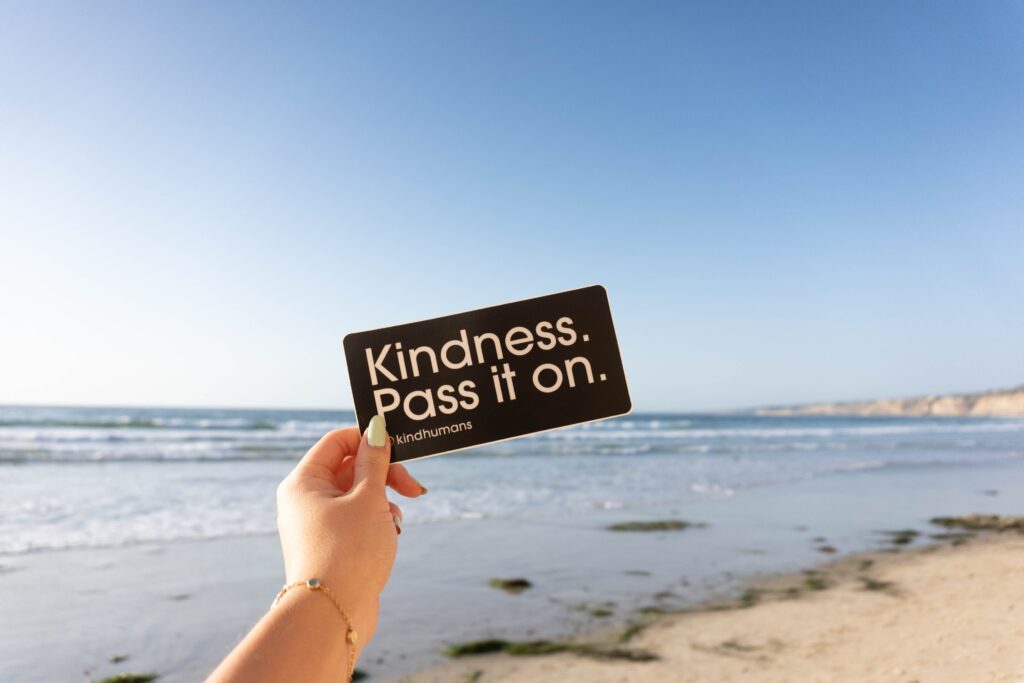 A person's hand holding a sign "Kindness, Pass it On" at a beach.