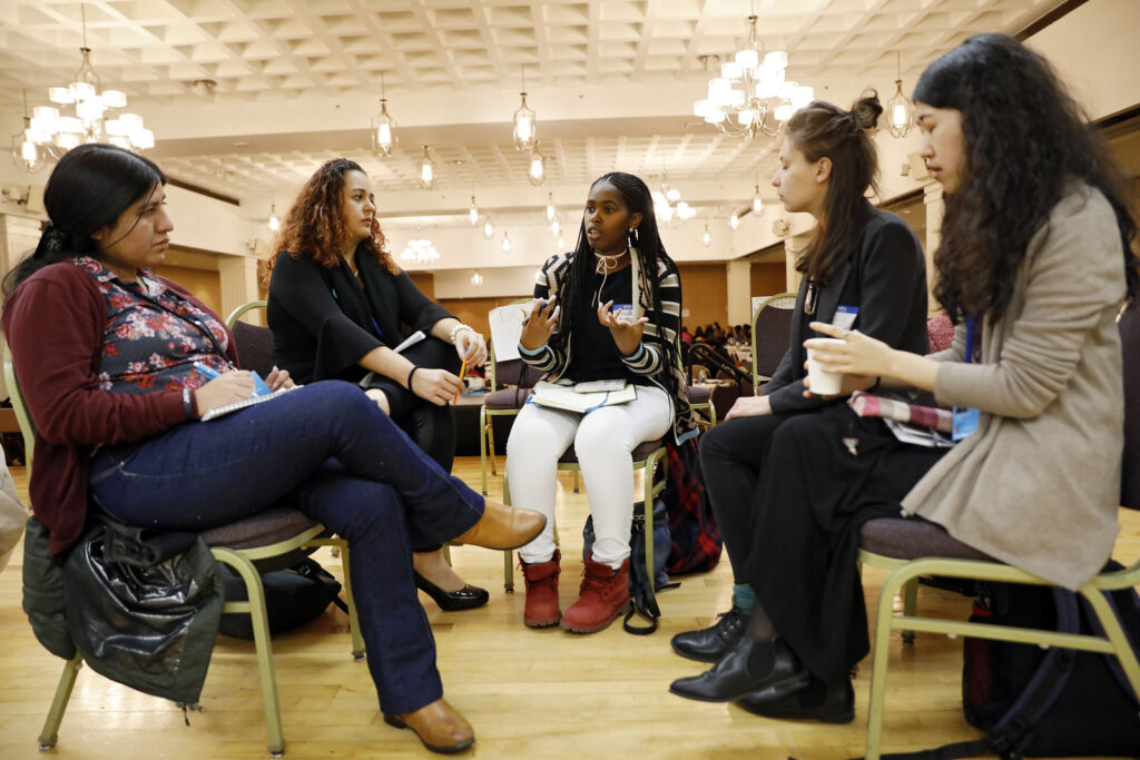 Five women from diverse backgrounds sitting in chairs discussing something. 