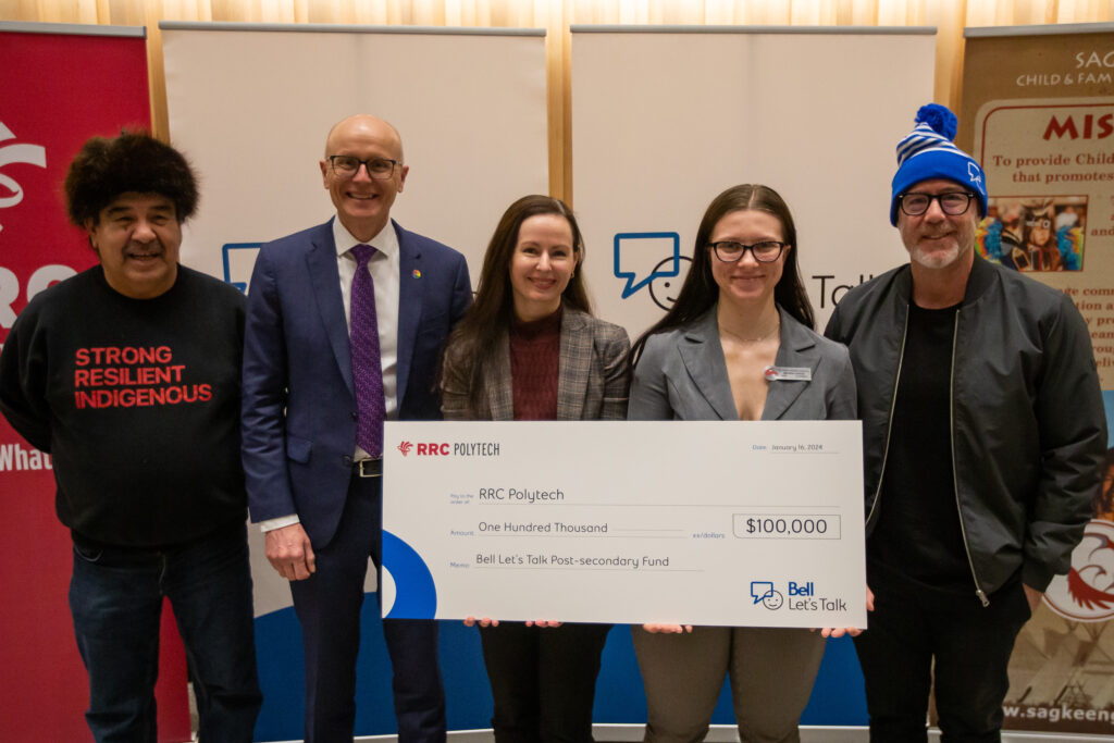 Five people stand in front of RRC Polytech and Bell Let's Talk banners, holding a giant cheque from Bell to RRC Polytech for one $100,000.