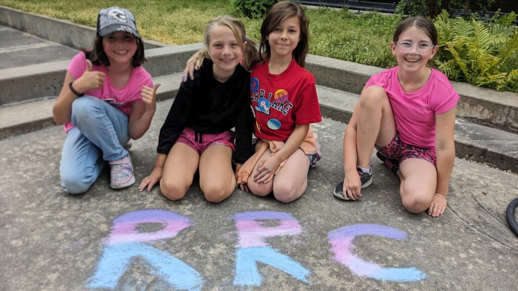 Four young campers kneeling behind pink and purple sidewalk chalk art that reads RRC