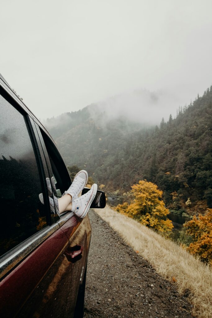 Side of car on the road with person's feet sticking out, driving through the mountains and fall coloured trees.