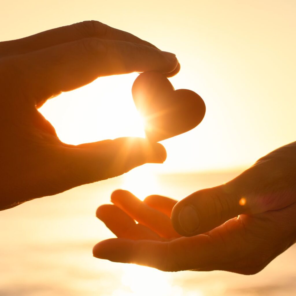two hand silhouettes one giving a heart shape rock to another with sun behind.