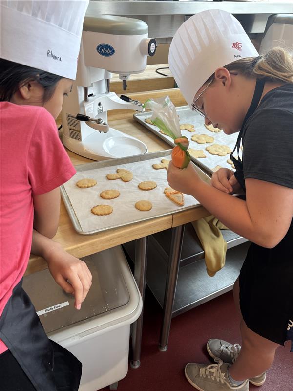 Two campers are decorating their freshly baked cookies using orange frosting