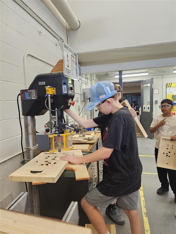 A camper is working on his operation style board while using a drill press