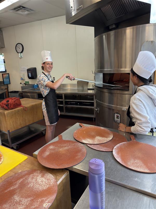 A camper is smiling at the camera while putting his pizza in the large pizza oven, another camper is standing by watching