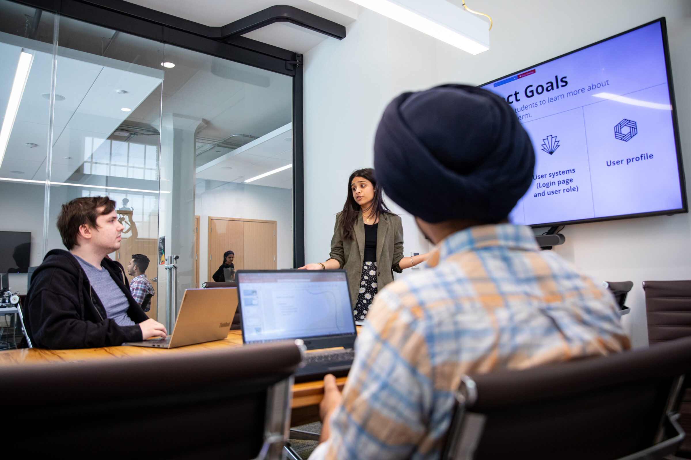 Two IT students sit at a table with laptops open in front of them while a third student stands at a smart board that says "Project Goals".
