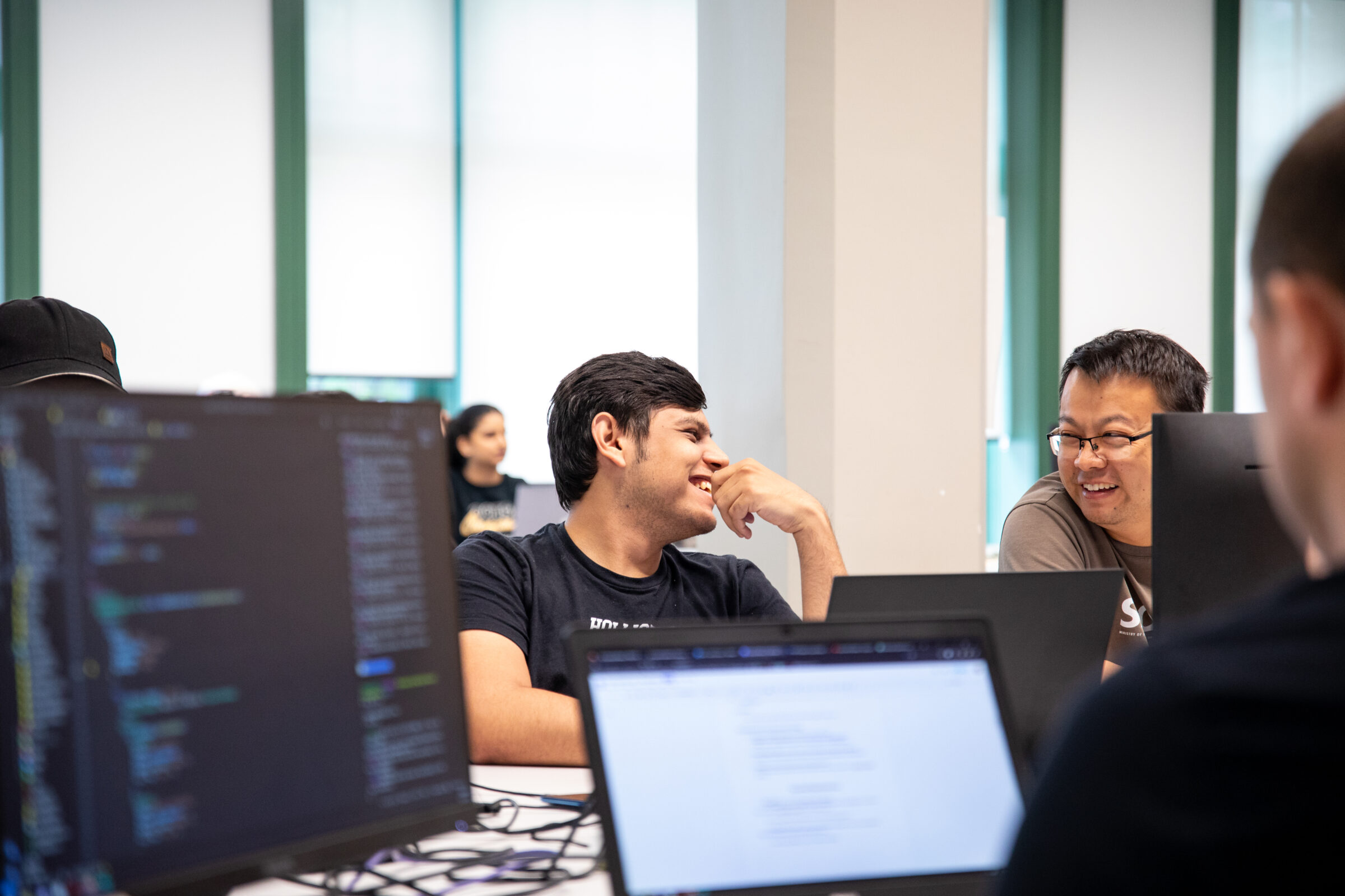 Foreground contains two monitors containing code. In the background, two students are looking at each other with smiles on their faces.