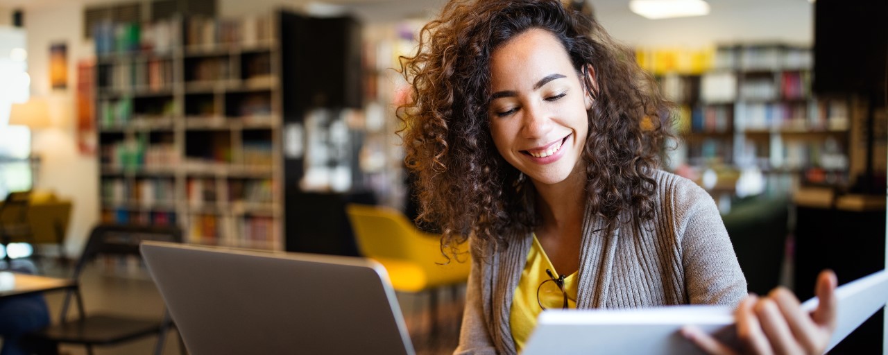 Female student in a library with a book in one hand with her laptop on the desk. 