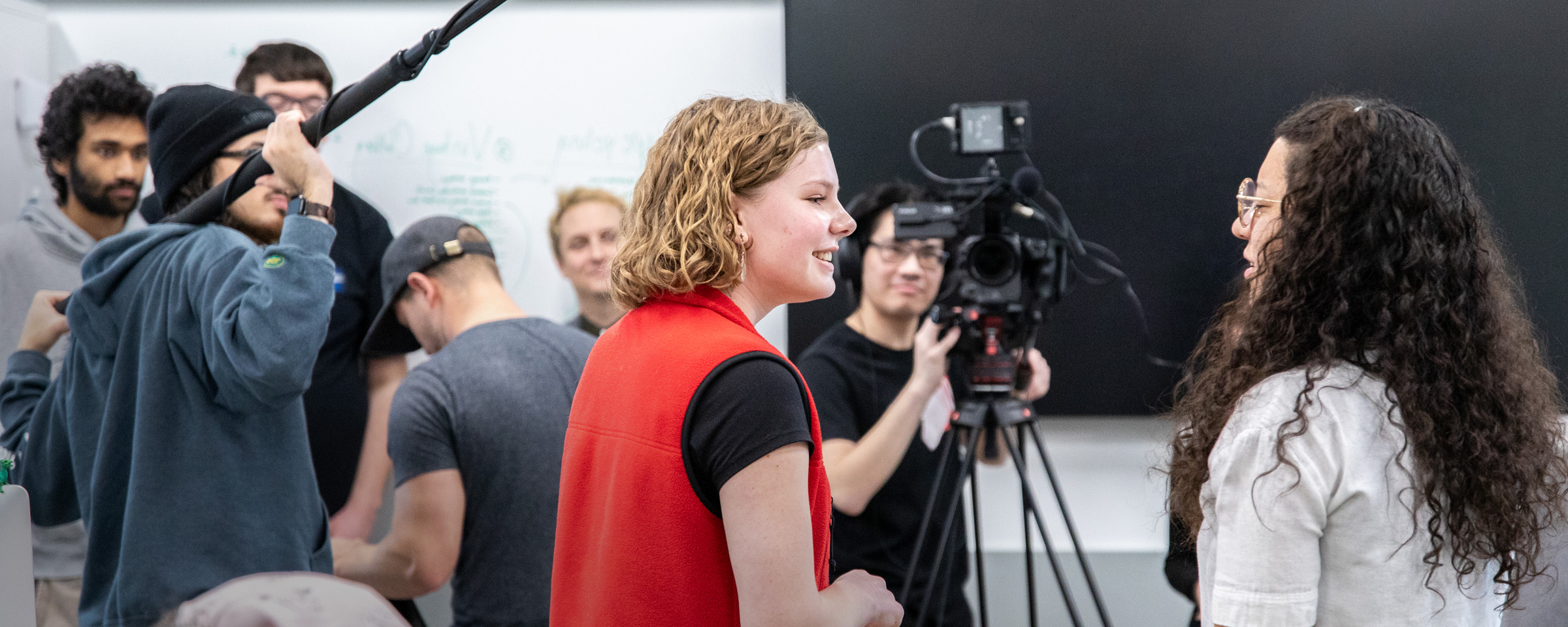 Two female students conduct a mock television interview while a male student shoots video and another operates a boom mic.