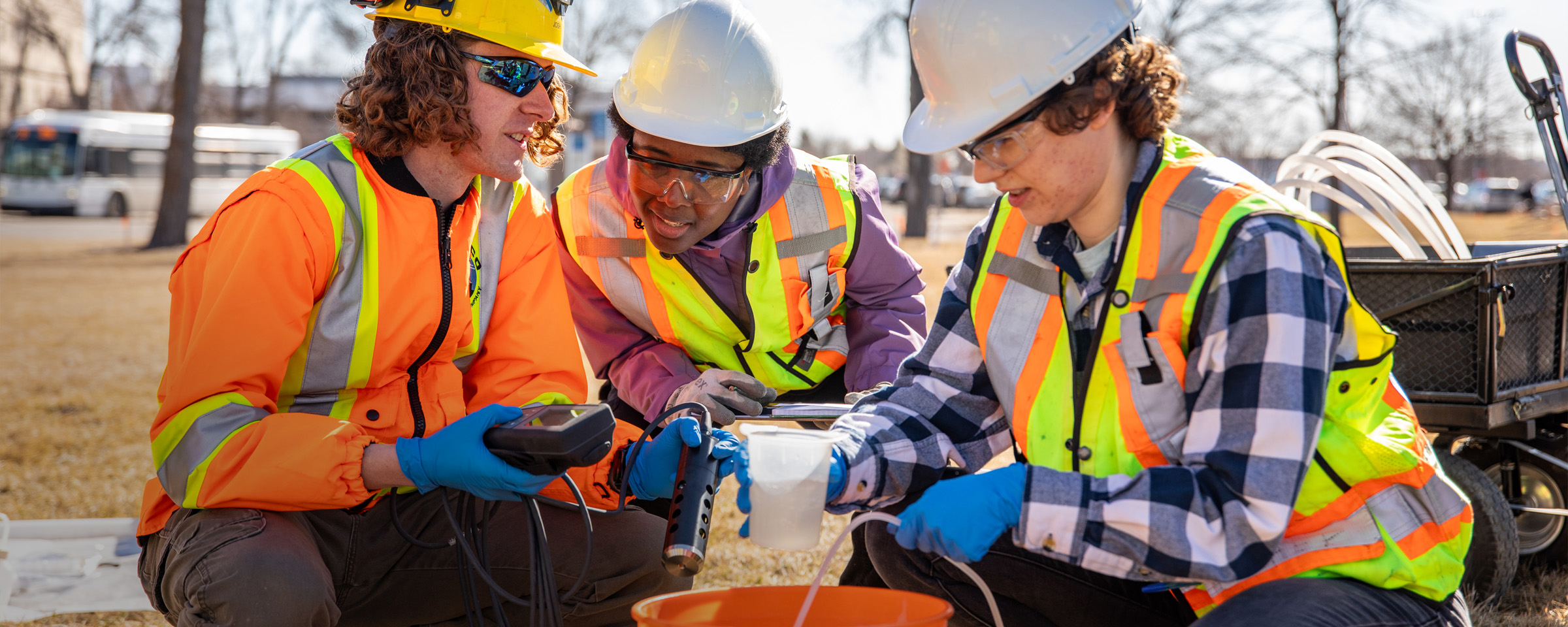 Trio of engineering students wearing safety vests, kneeling around a pail while testing water quality outdoors