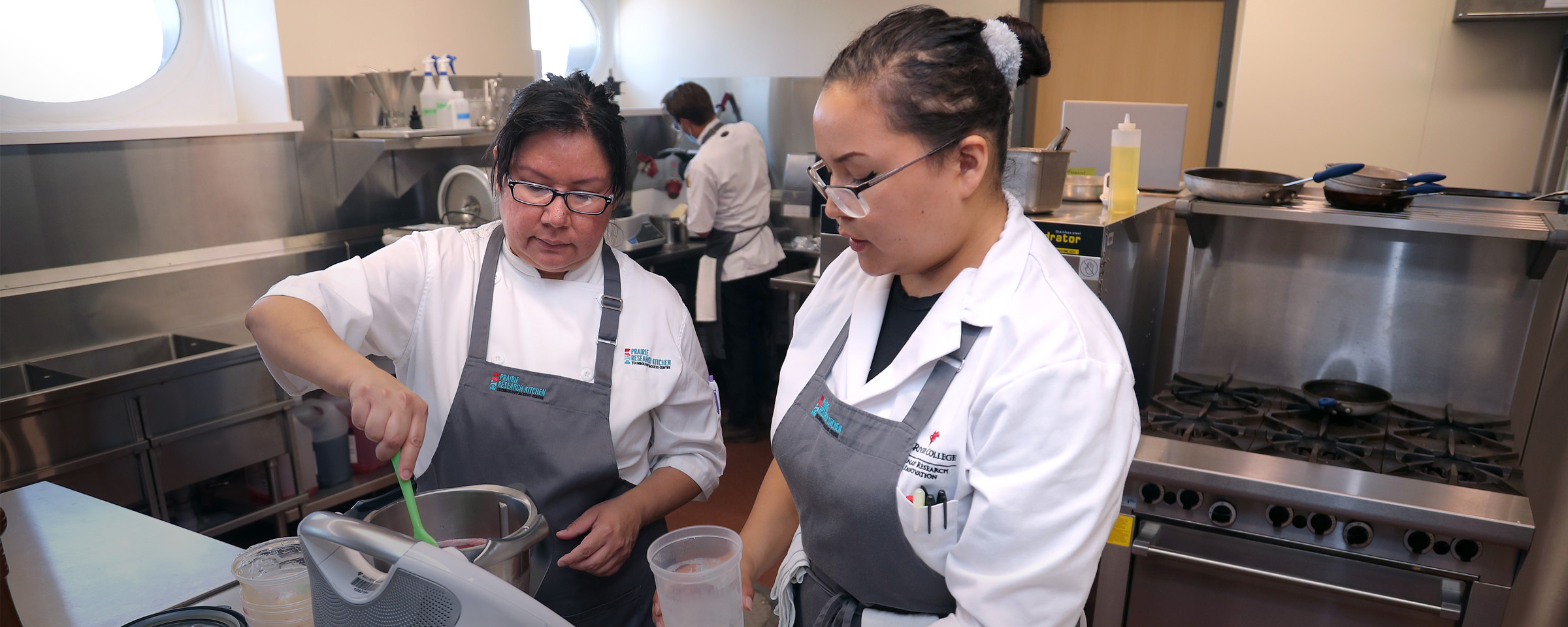 Two Indigenous women wearing chefs' smocks and aprons stirring ingredients inside RRC Polytech's Prairie Research Kitchen.