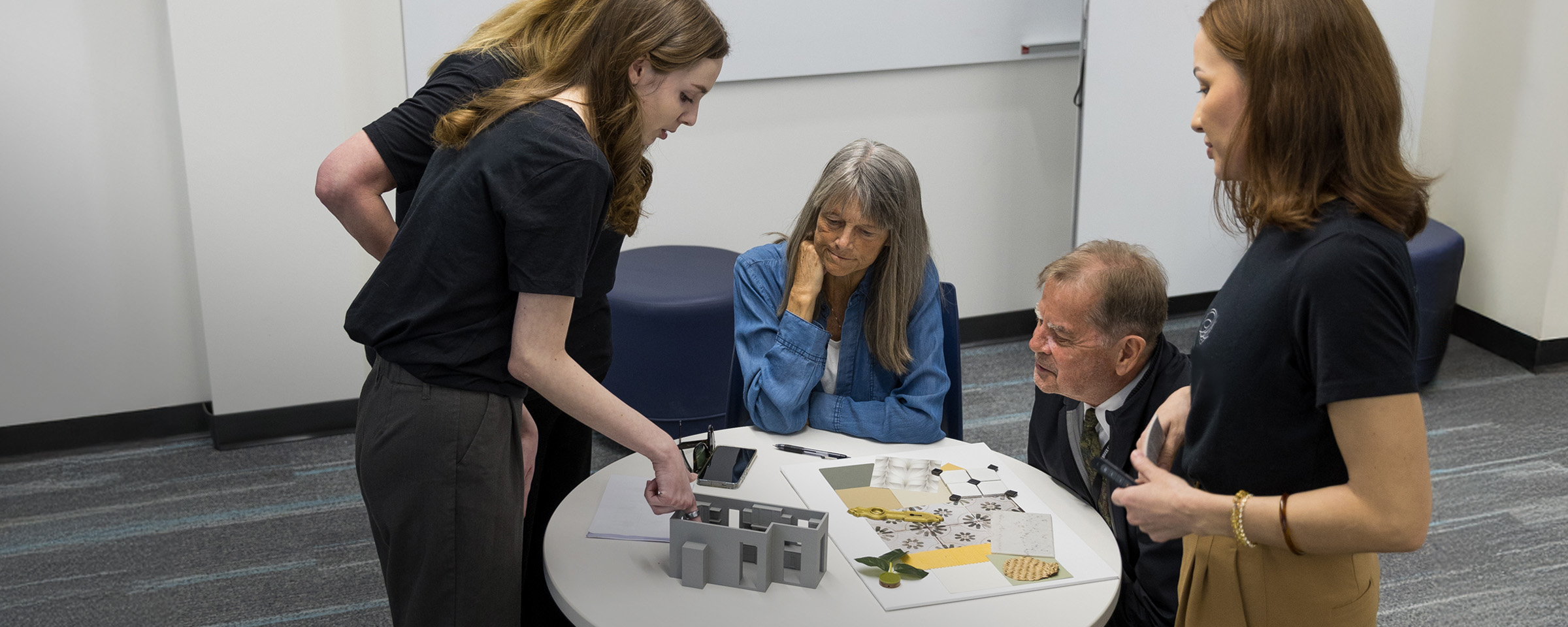 Three female students present an interior decorating mock-up display, while two clients, seated at a table, look on.
