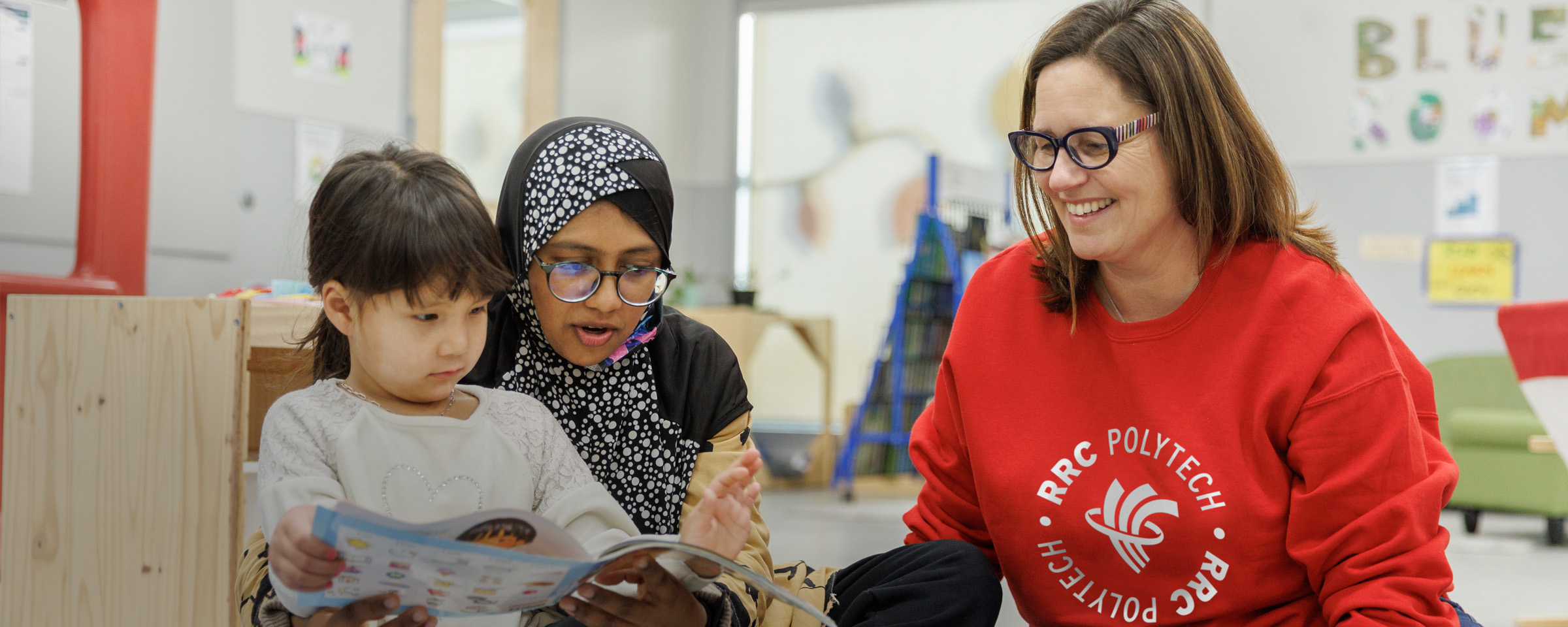 Female RRC Polytech staff member, seated, with adult female day care worker and young child reading a book.