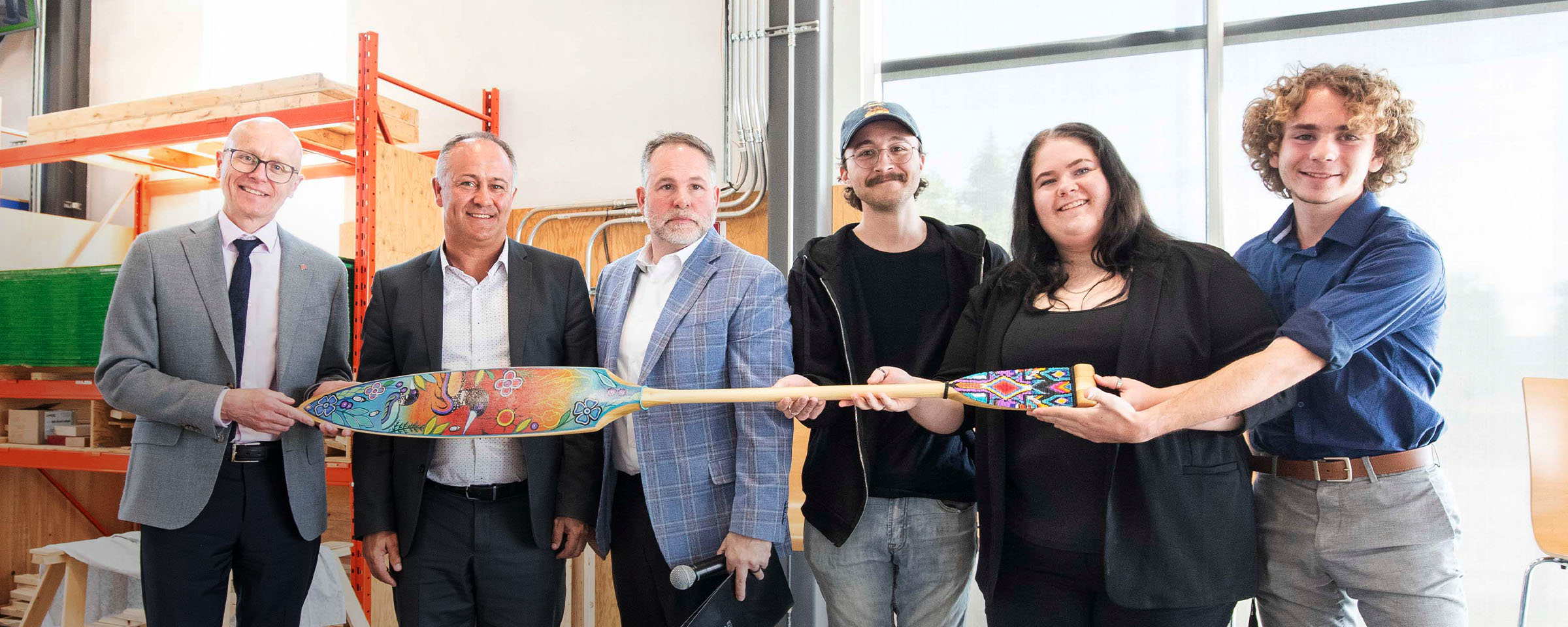 Row of six people, standing, holding a wooden paddle painted with Indigenous designs.