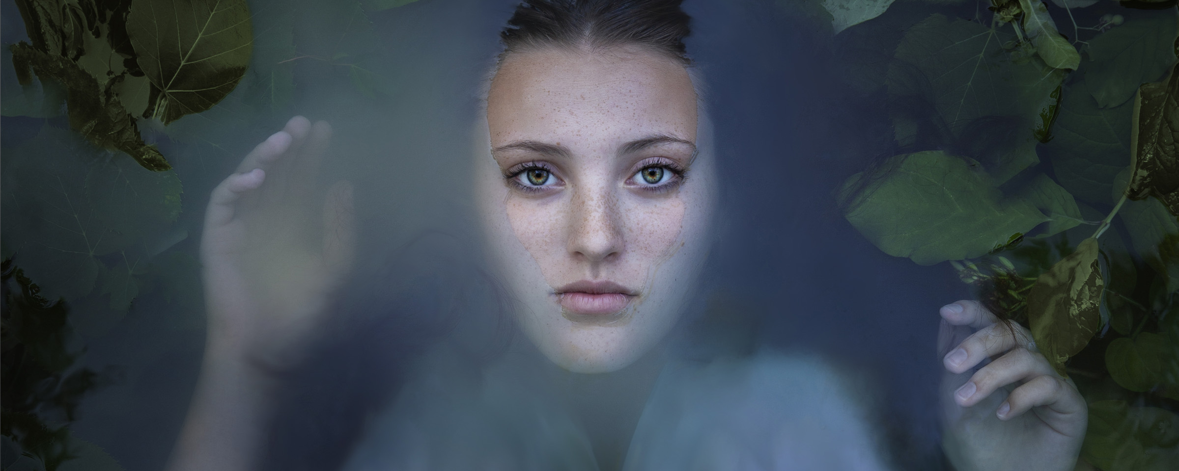 Artistic portrait of woman's face, half-submerged in water and framed by plant life.