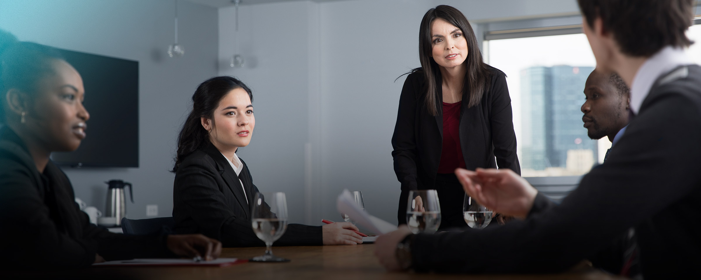 A woman in black business attire stands at a boardroom table, flanked by two seated men and two seated women.