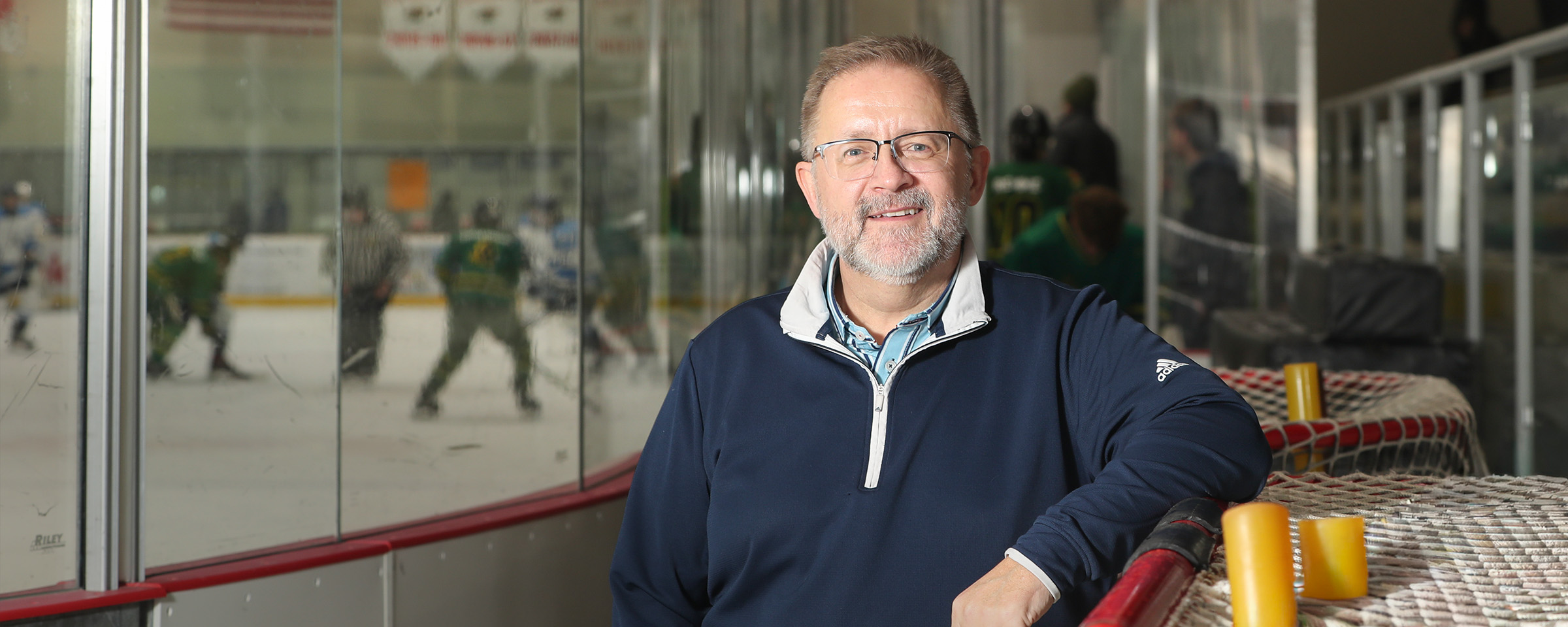 Trevor Buhnai, wearing blue half-zip pullover and glasses, stands at the boards of a hockey rink with players and ref on ice in the background.