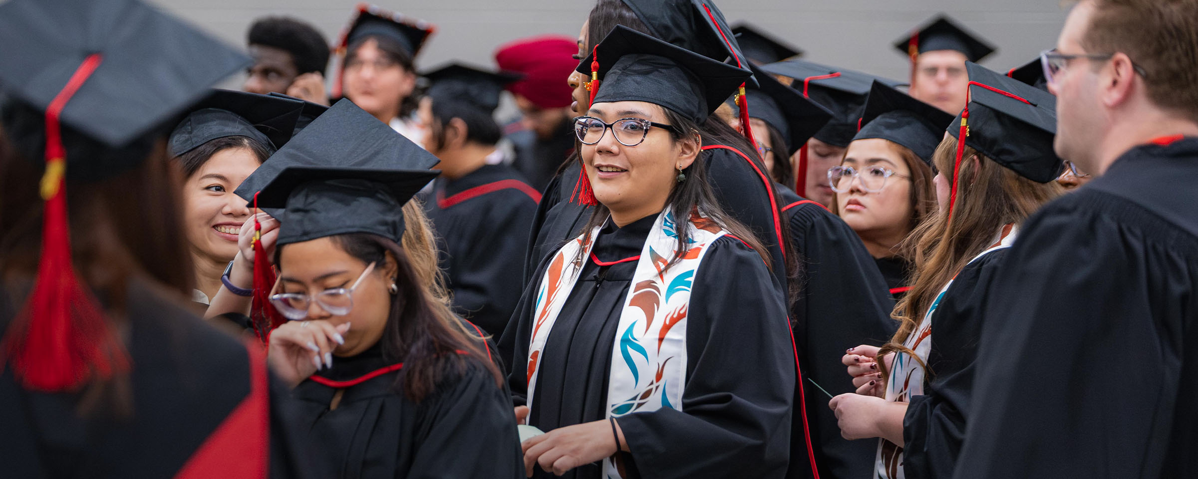 Female student, wearing graduation cap and gown and Indigenous stole, surrounded by other graduates at RRC Polytechnic convocation ceremony.