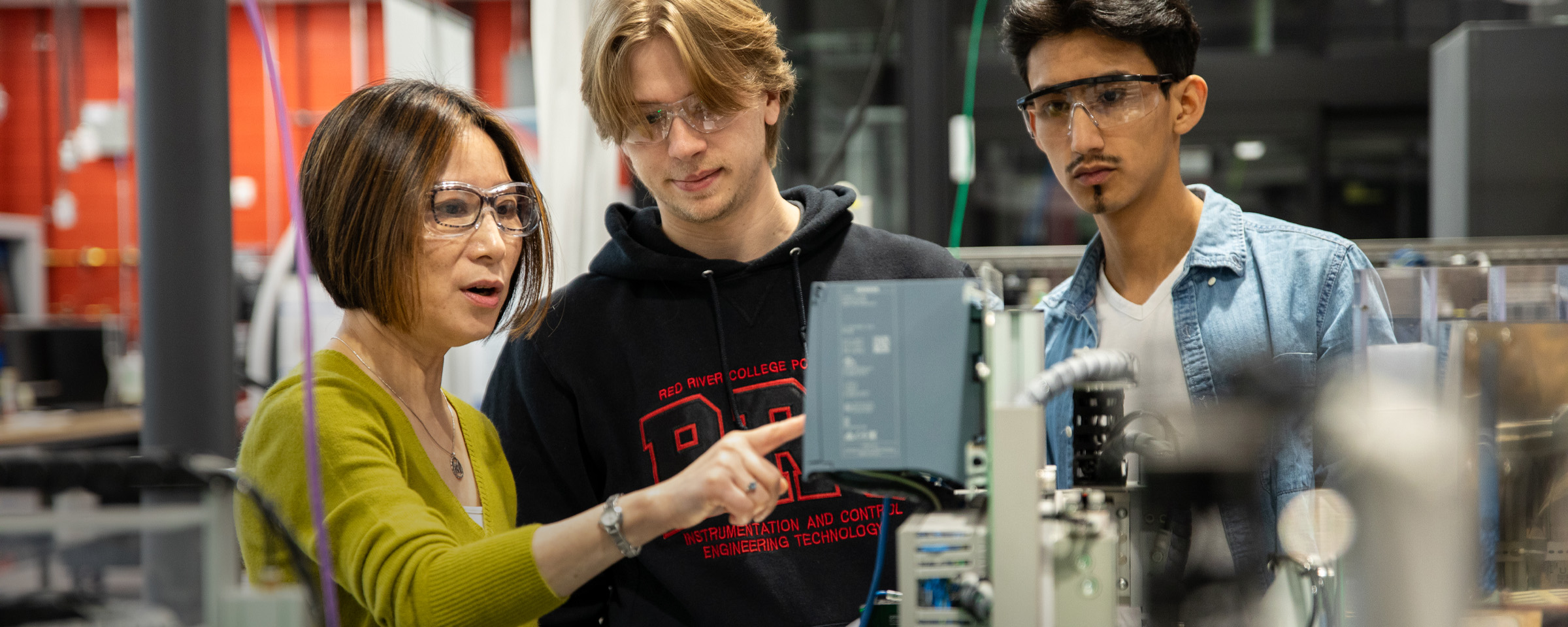 Female instructor programs manufacturing equipment as two make students look on.